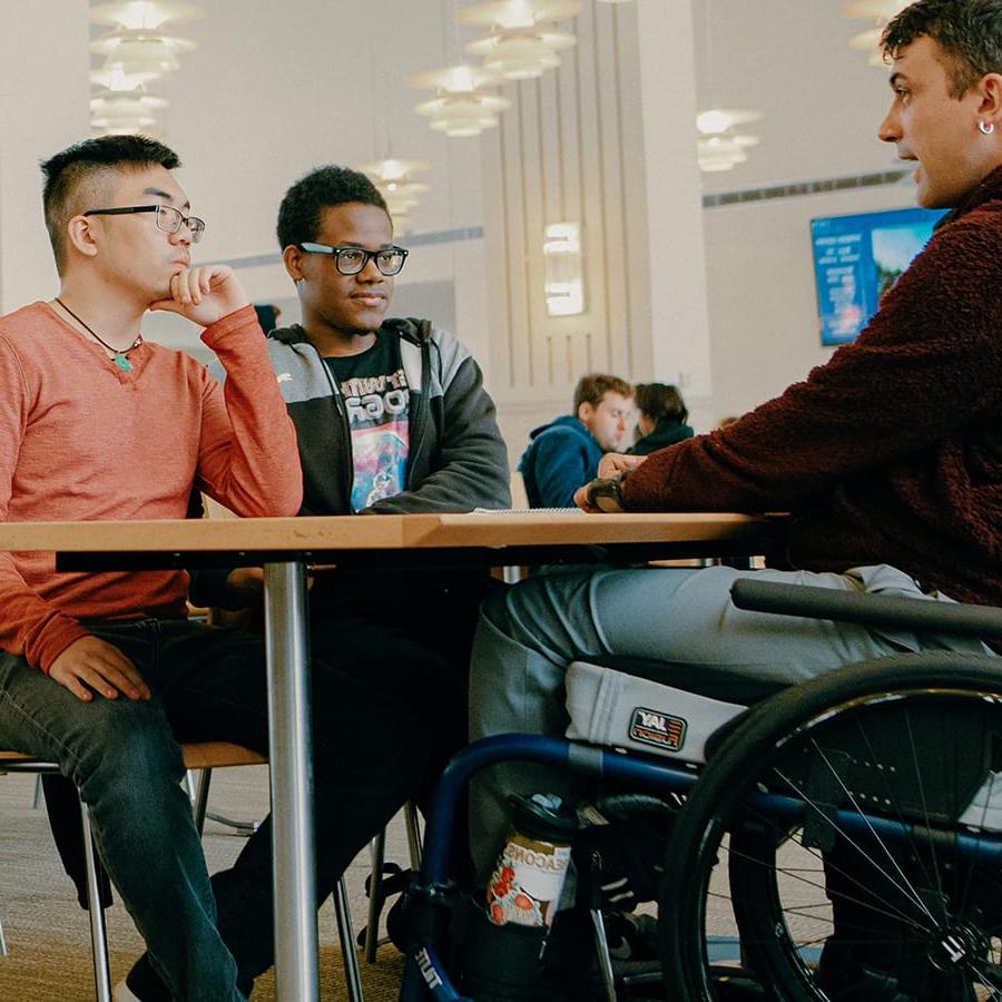 Stefan sits in wheelchair talks with two students in cafeteria.